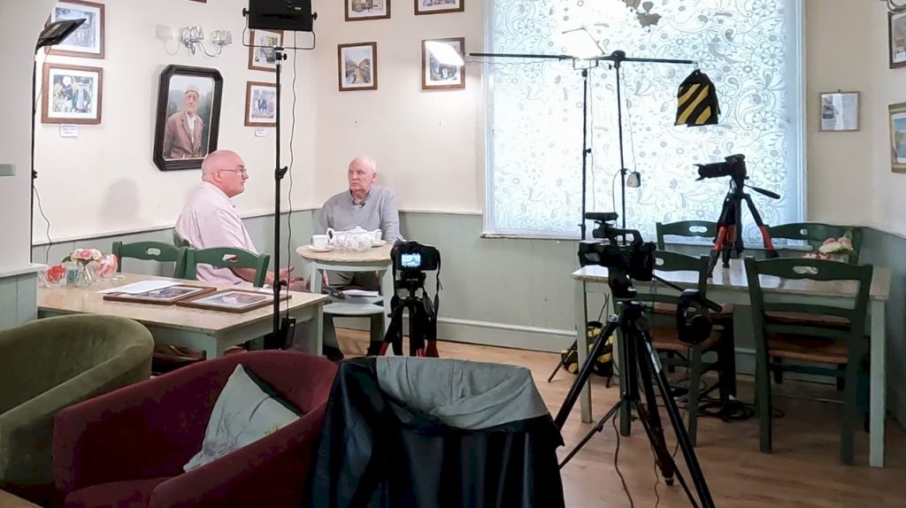 Wide view of interior of The Wrinkled Stocking Tearoom, showing Leigh and Terry at a table with a pot of tea, showing the video cameras and lights used for the video shoot.